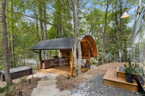 Una cabaña de madera con un porche cubierto, mobiliario exterior y zona de barbacoa, rodeada de árboles.