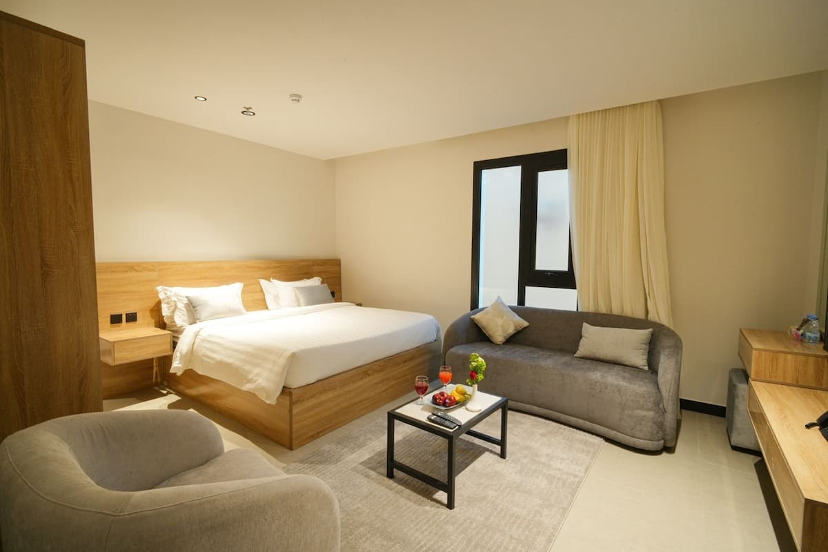 A modern hotel room with a large bed, a sofa, a chair, a small table with fruit and drinks, and a window with curtains.