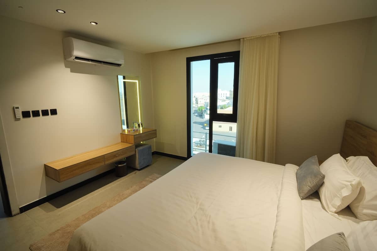 A modern hotel room with a large bed, a built-in desk, a mirror, and a view of the city through a large window.
