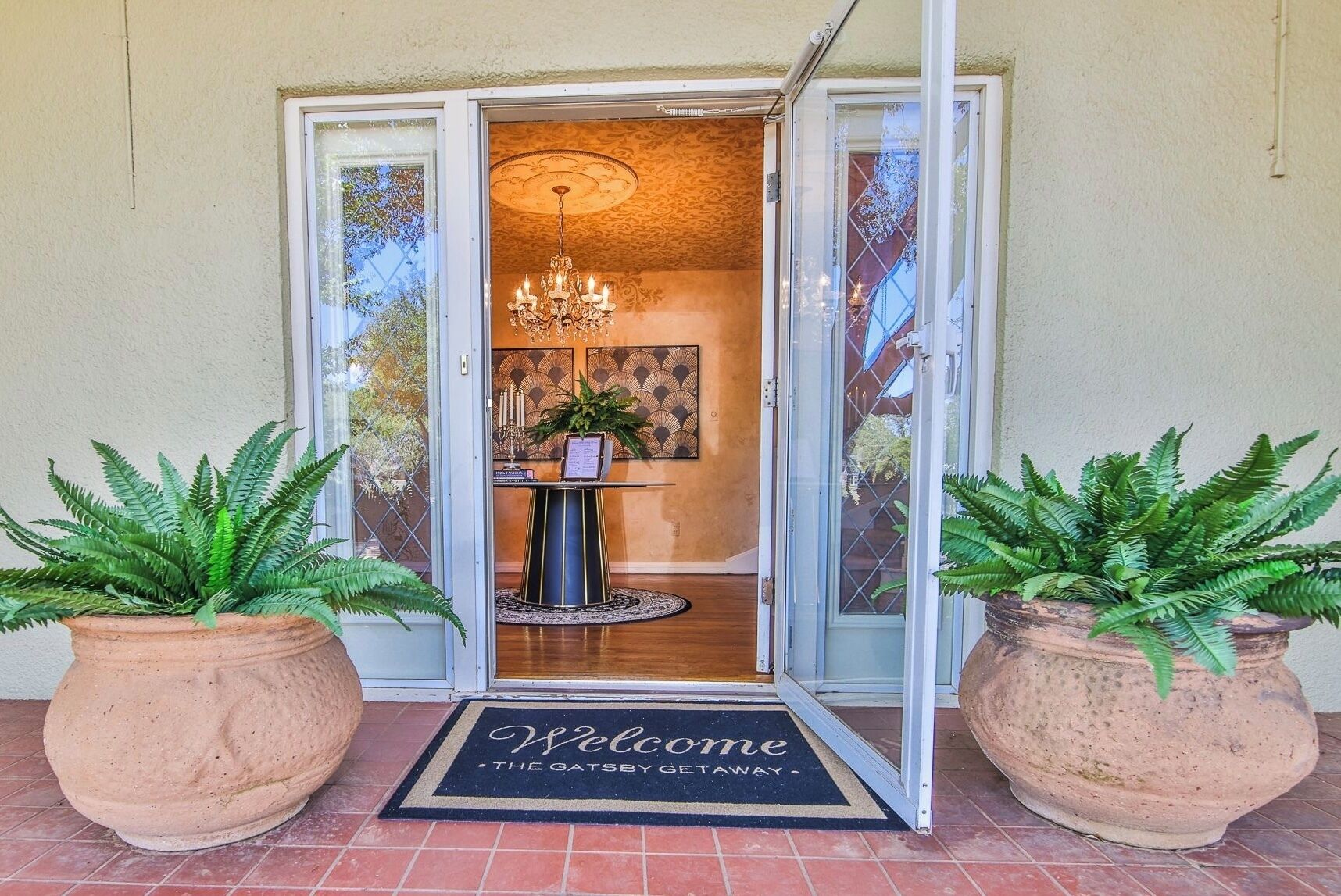 A hotel entrance with a glass door, a welcome mat, and two large potted plants.