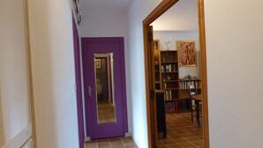 Käytävällä on violetti ovi, joka johtaa kylpyhuoneeseen, siellä on myös puinen kirjahylly ja lattia on laatoitettu.