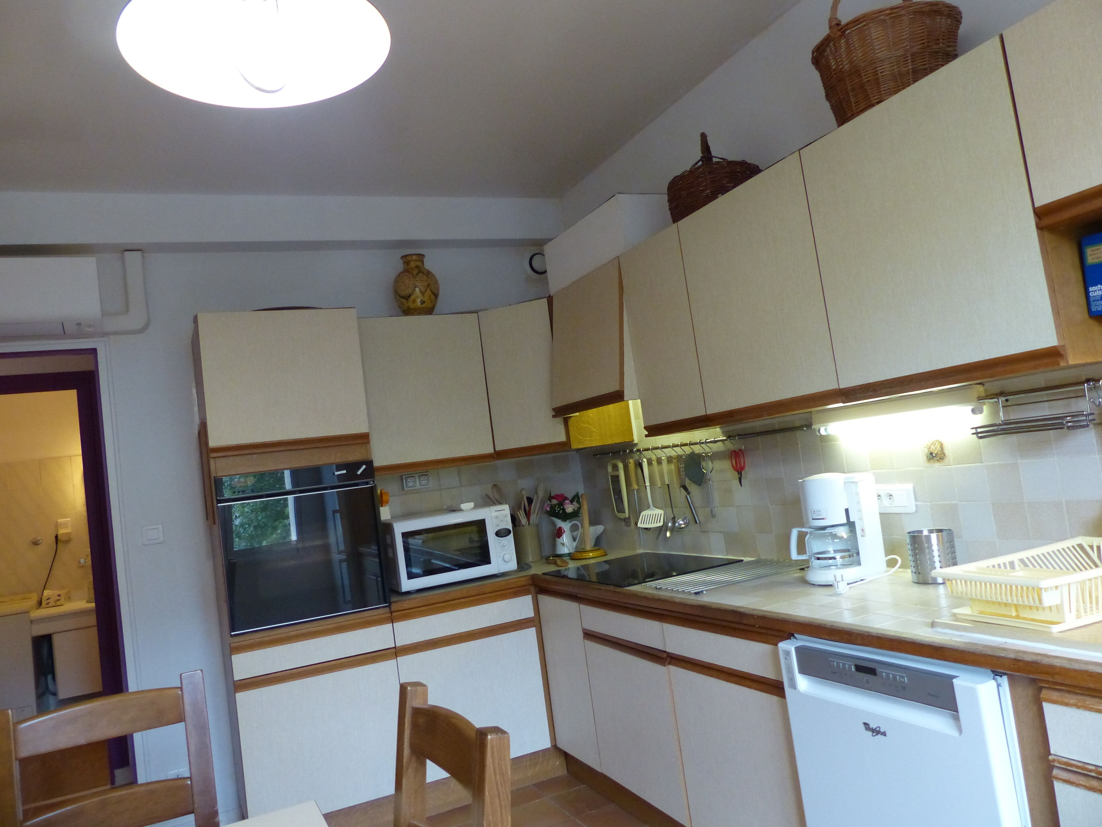 Cocina compacta con muebles blancos, microondas, estufa y lavavajillas. Hay una silla de madera y una mesita.