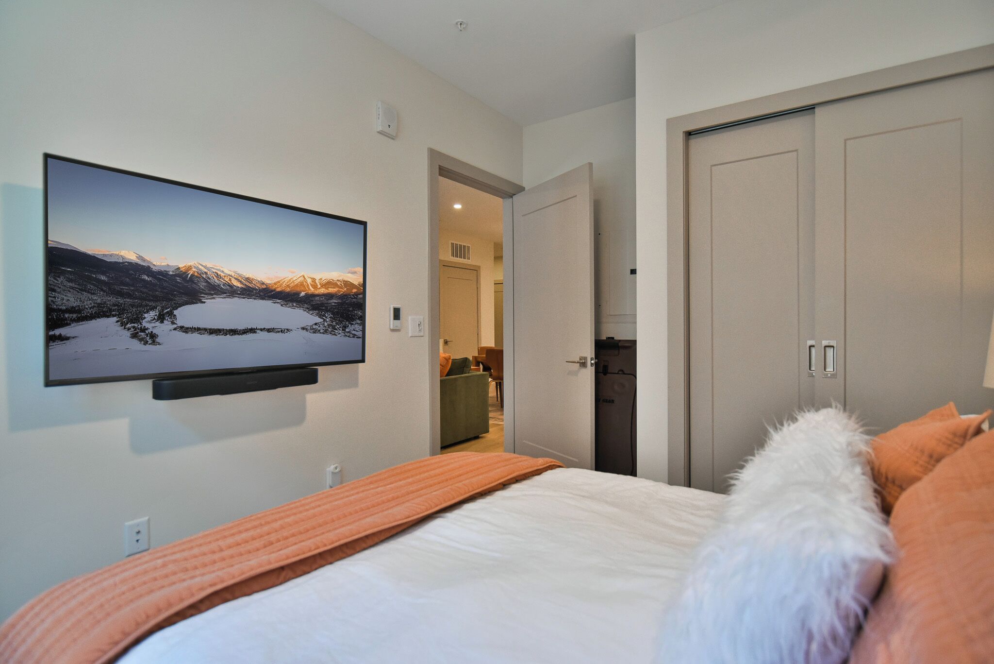 Une chambre à coucher comprenant un lit, un téléviseur fixé au mur qui diffuse un paysage pittoresque, une armoire et une porte donnant sur une autre pièce.