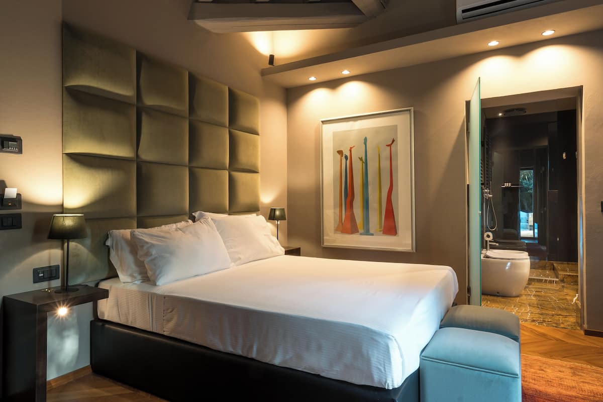 Une chambre d’hôtel moderne avec un grand lit, une tête de lit au design original, une table de chevet avec une lampe et un tableau au mur.