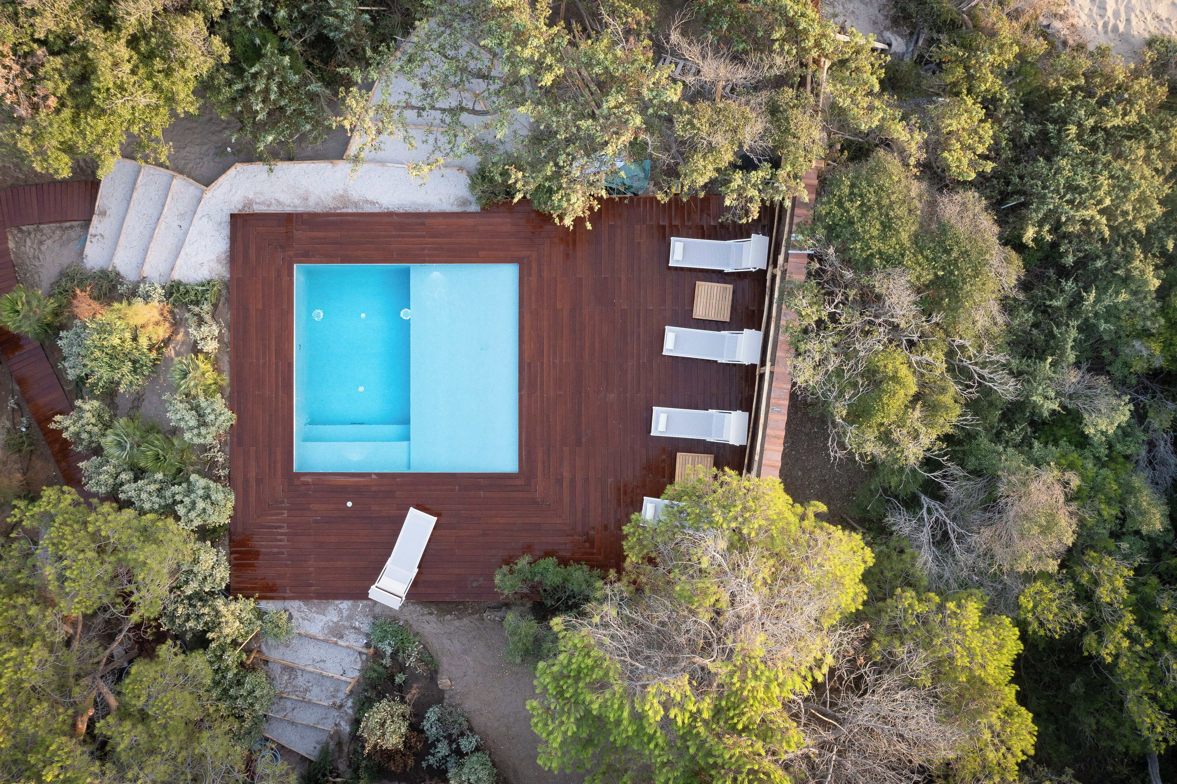 An aerial view of a rectangular swimming pool with a wooden deck, surrounded by dense foliage.