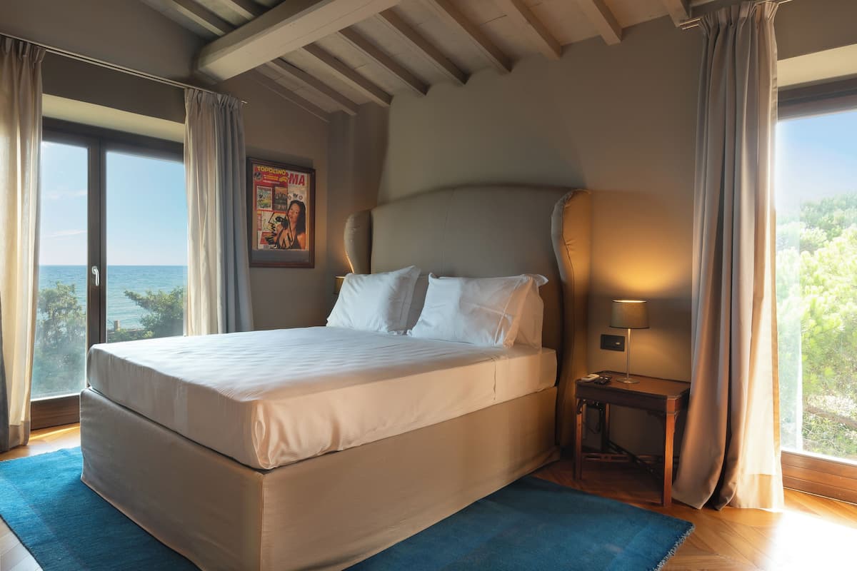 Un grand lit avec du linge de lit blanc, une table de chevet avec une lampe et une vue sur la mer par de grandes fenêtres.