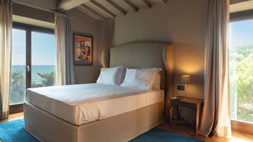 A large bed with white linens, a nightstand with a lamp, and a view of the sea through large windows.