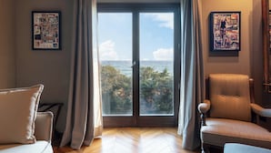 A room with a large sliding glass door leading to a balcony with a view of the sea. There are two framed pictures on the walls, a chair, and a small table.