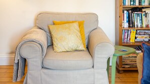 Un sillón de color gris claro con un cojín amarillo, una mesita auxiliar verde pequeña y una estantería de madera llena de libros y DVD.