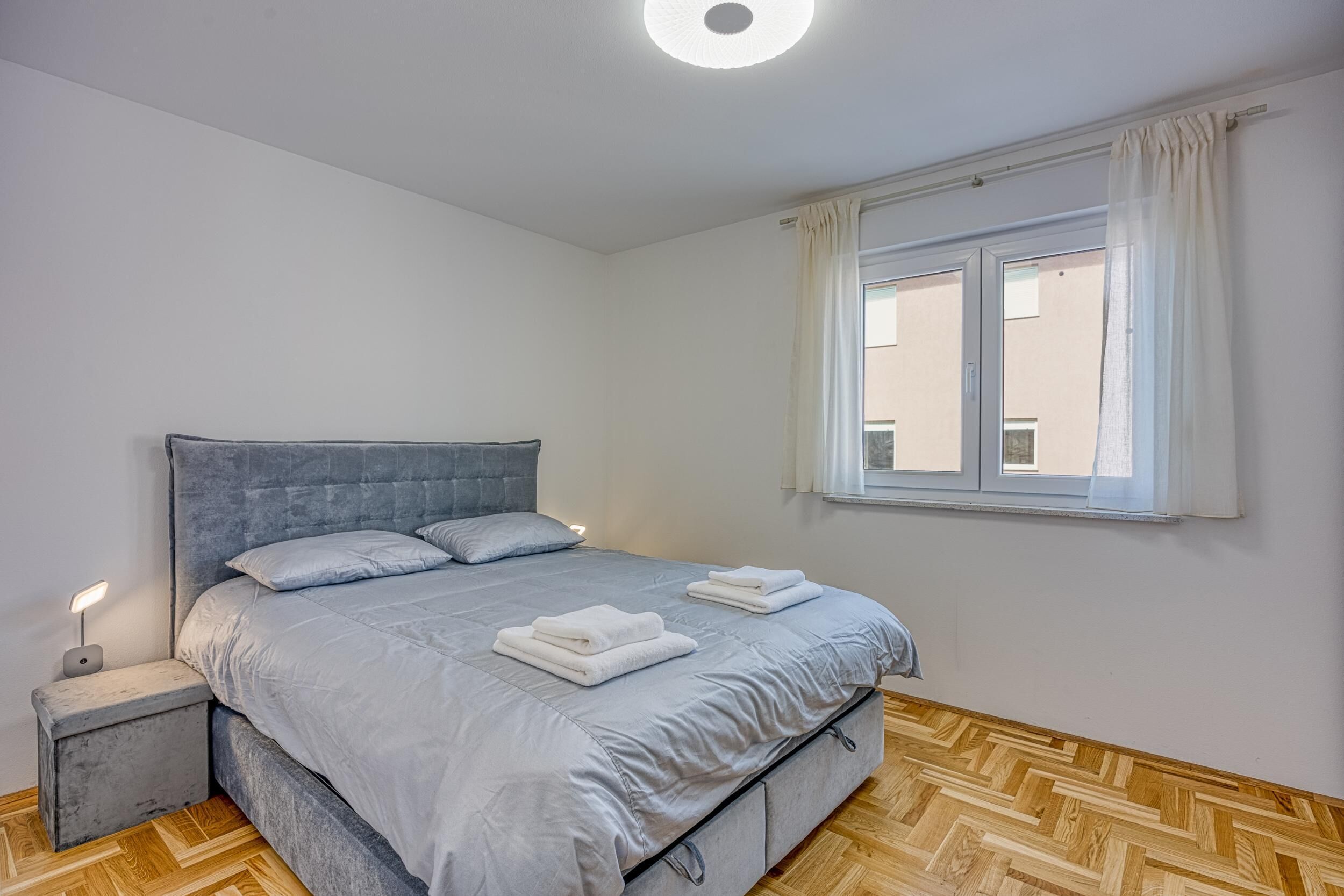 Et soveværelse med en grå seng, hvide puder, et natbord med lampe og et vindue med gardiner.