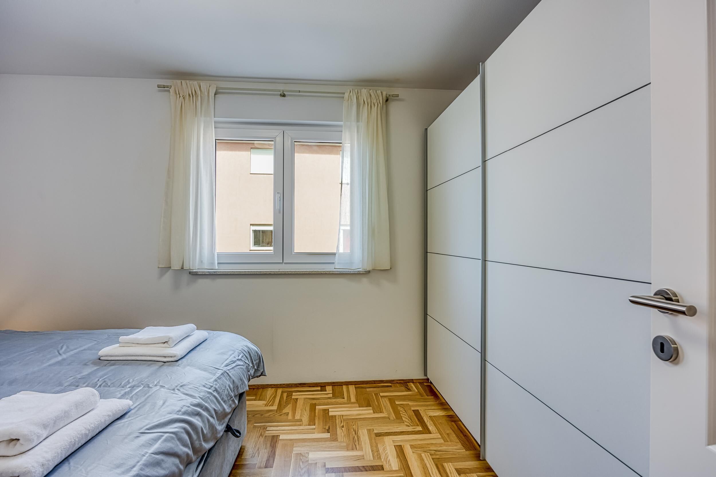 Un dormitorio con cama, armario, ventana con cortinas y suelo con diseño en espiga.
