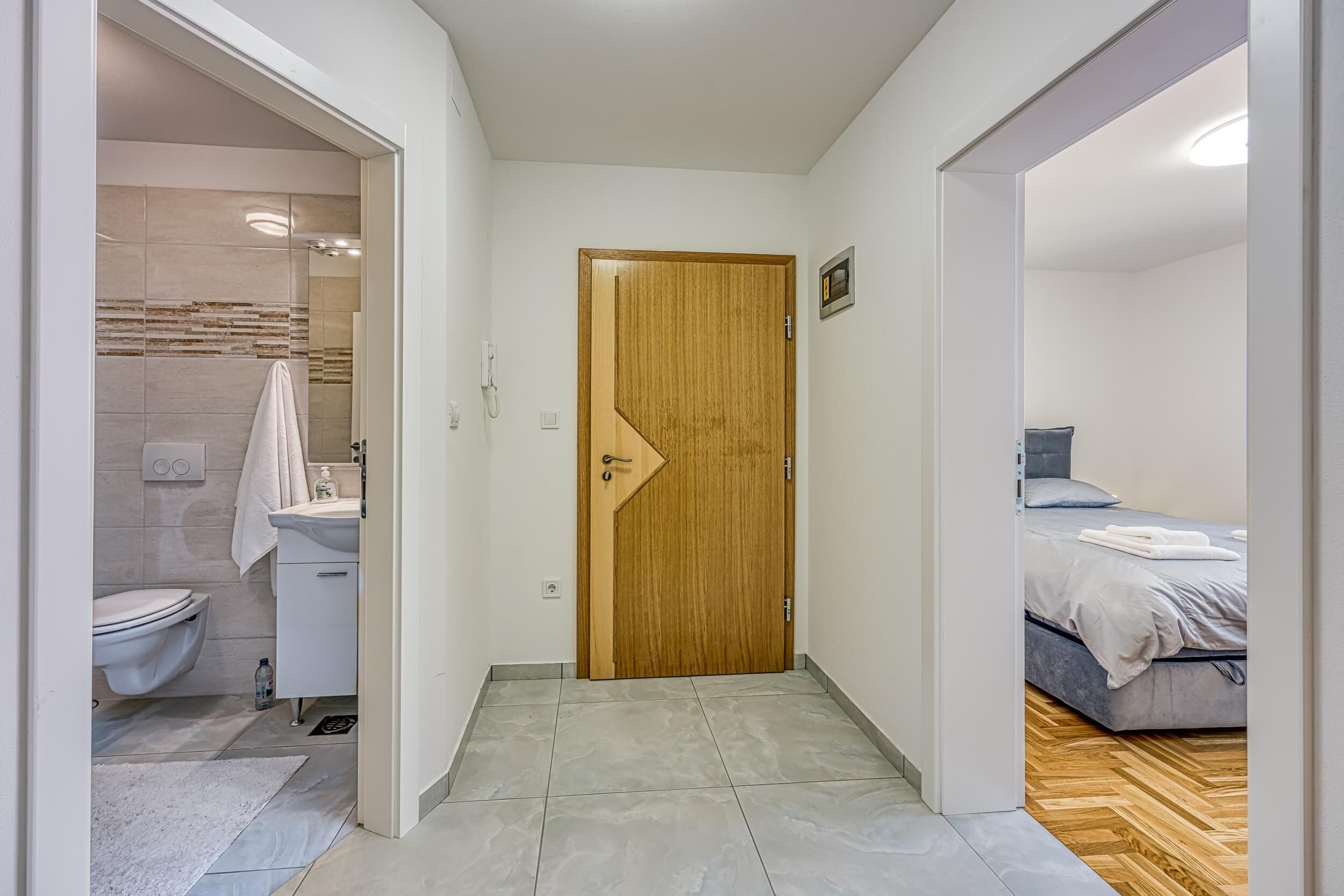 Een gang die leidt naar een badkamer met toilet, wastafel en douche. In de aangrenzende kamer staat een bed met matras en kussens.