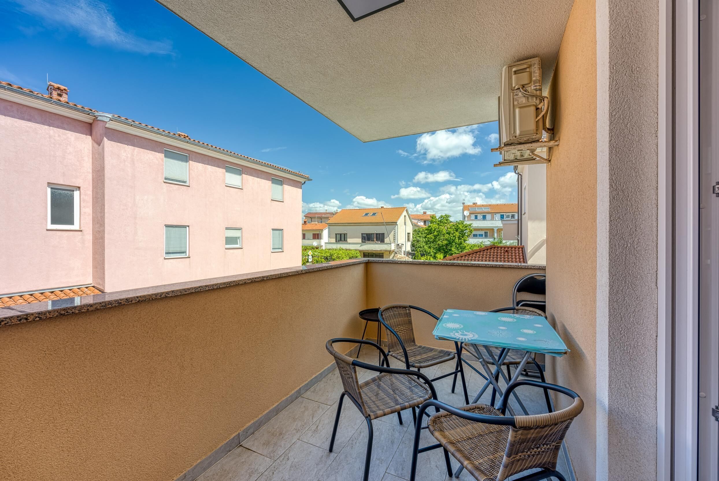 A balcony with wicker chairs and a table, overlooking neighboring buildings and a clear sky.