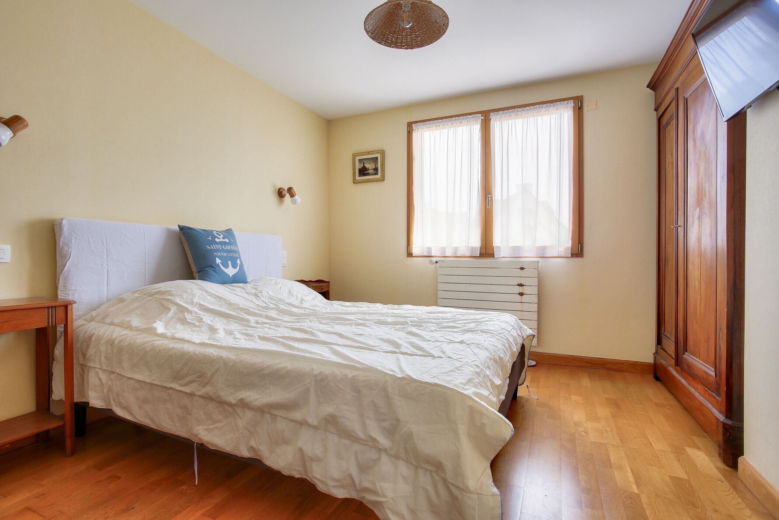 Un dormitorio con cama, suelo de madera, ventana con cortinas, armario y mesita de noche.