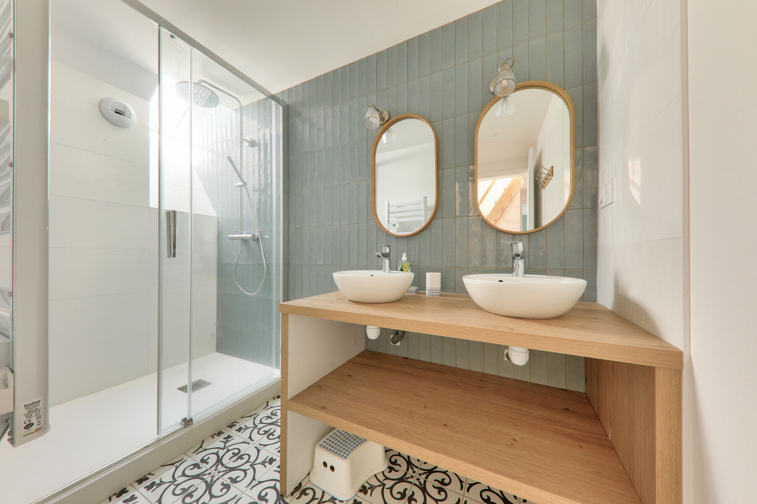 A modern bathroom with a glass shower enclosure, two sinks with round mirrors, and patterned floor tiles.