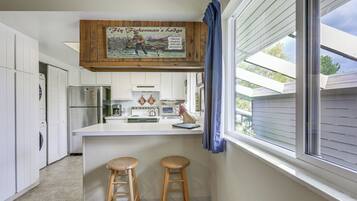 Una cocina con un letrero de madera sobre la encimera, una ventana con persianas y dos taburetes.