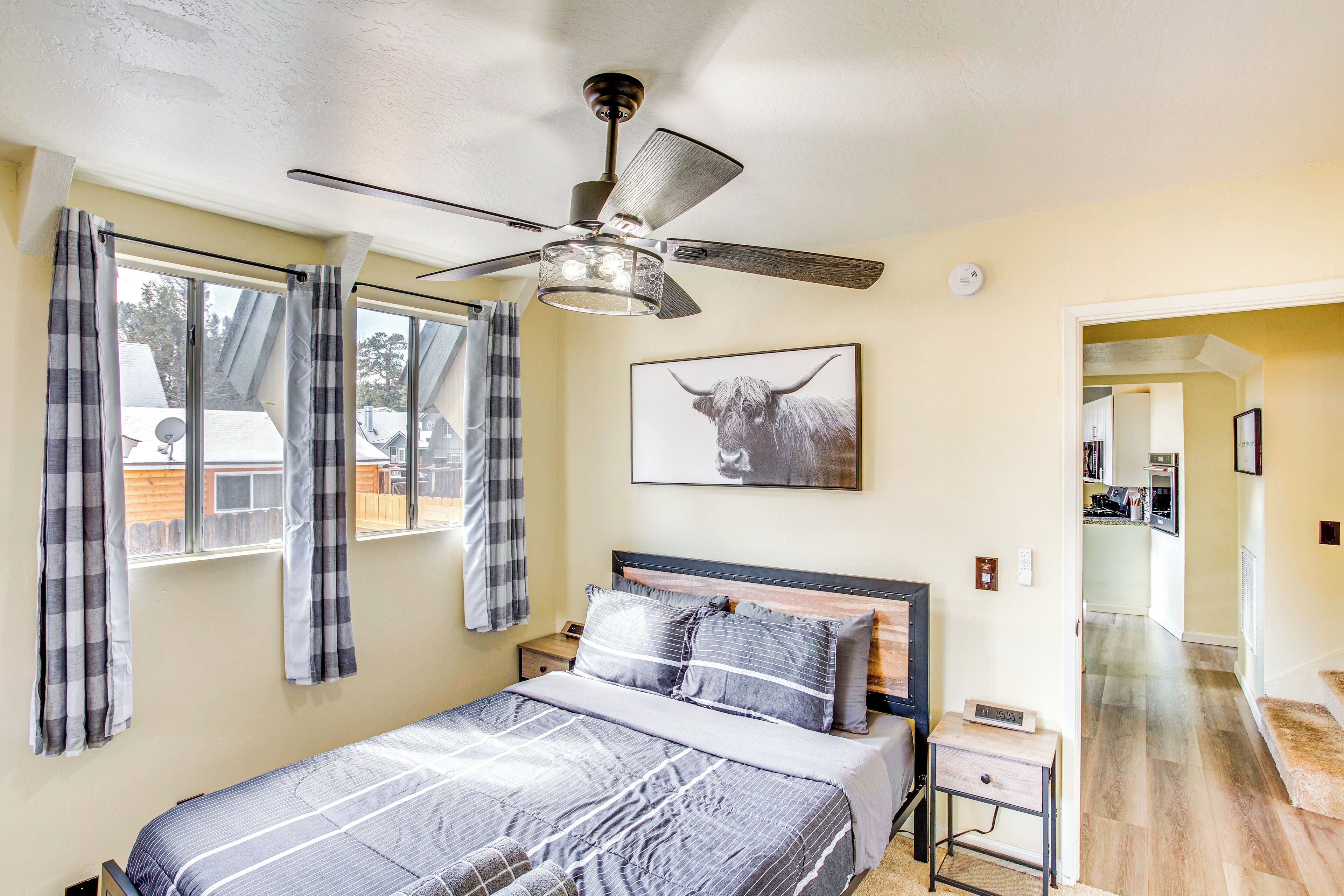 A bedroom with a bed, a ceiling fan, a window with curtains, a framed picture on the wall, and a hallway leading to a kitchen.