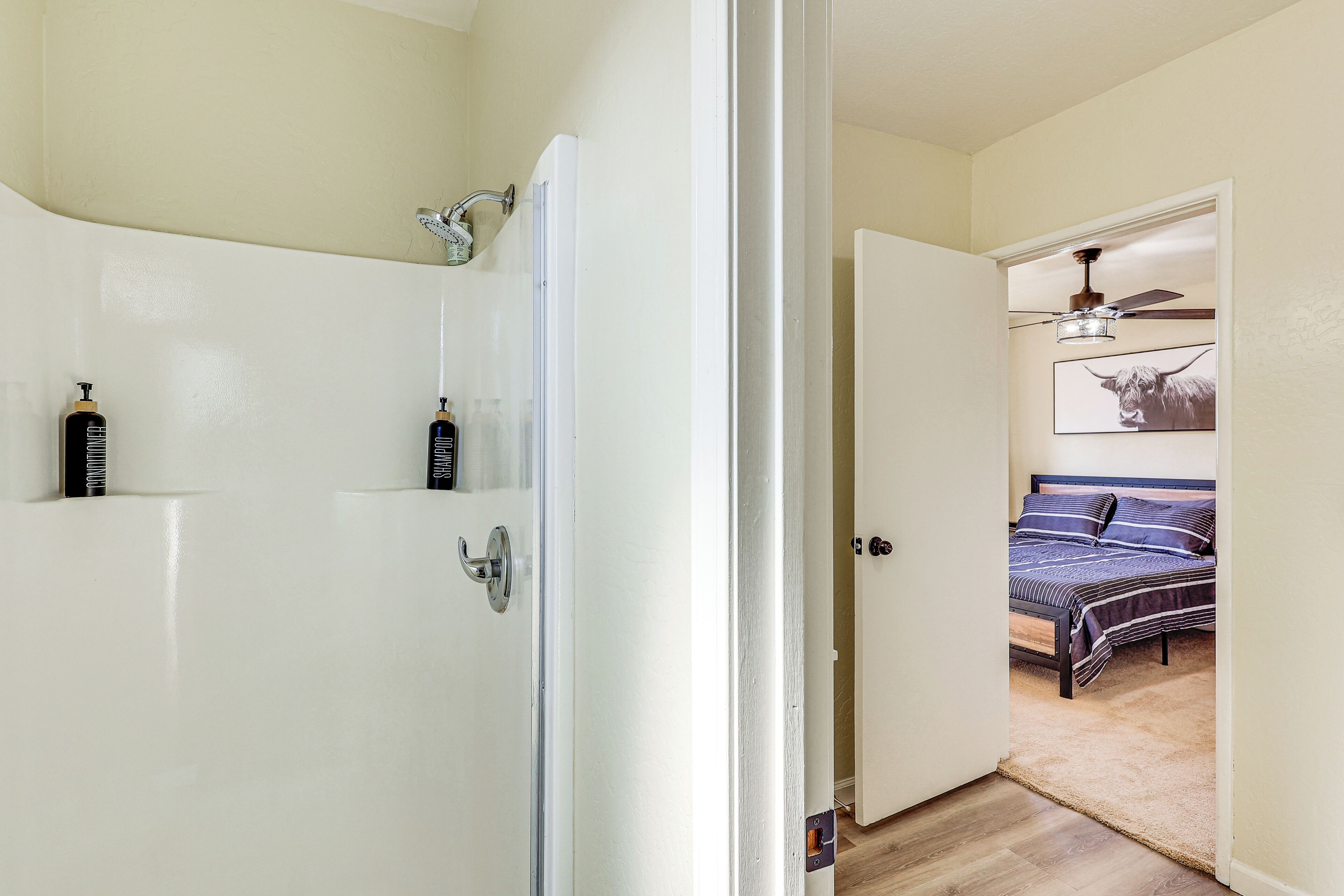 A bathroom with a bathtub, a shower, and a toilet. There is a bed with a headboard and a ceiling fan in the adjacent room.