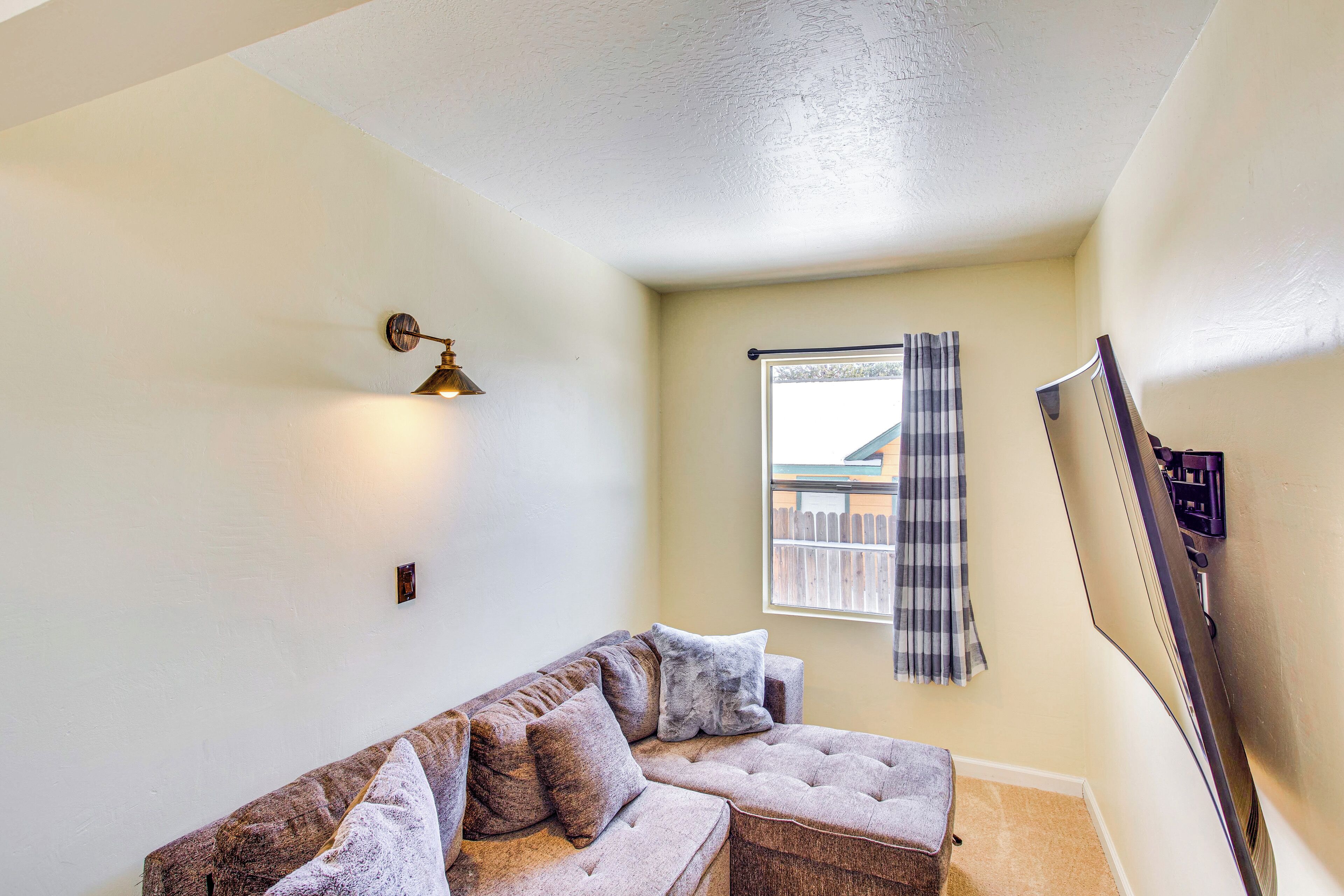 A corner room with a sectional sofa, a wall-mounted TV, a window with a view of a fence, and a ceiling light fixture.