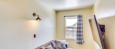A corner room with a sectional sofa, a wall-mounted TV, a window with a view of a fence, and a ceiling light fixture.