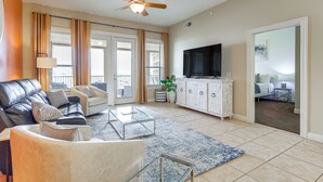 A spacious living room with a leather sofa, a glass coffee table, and a television. There is a ceiling fan, a large window with curtains, and a white cabinet. - Lake LBJ Condo w/ Private Balcony, Lake Views (Horseshoe Bay)