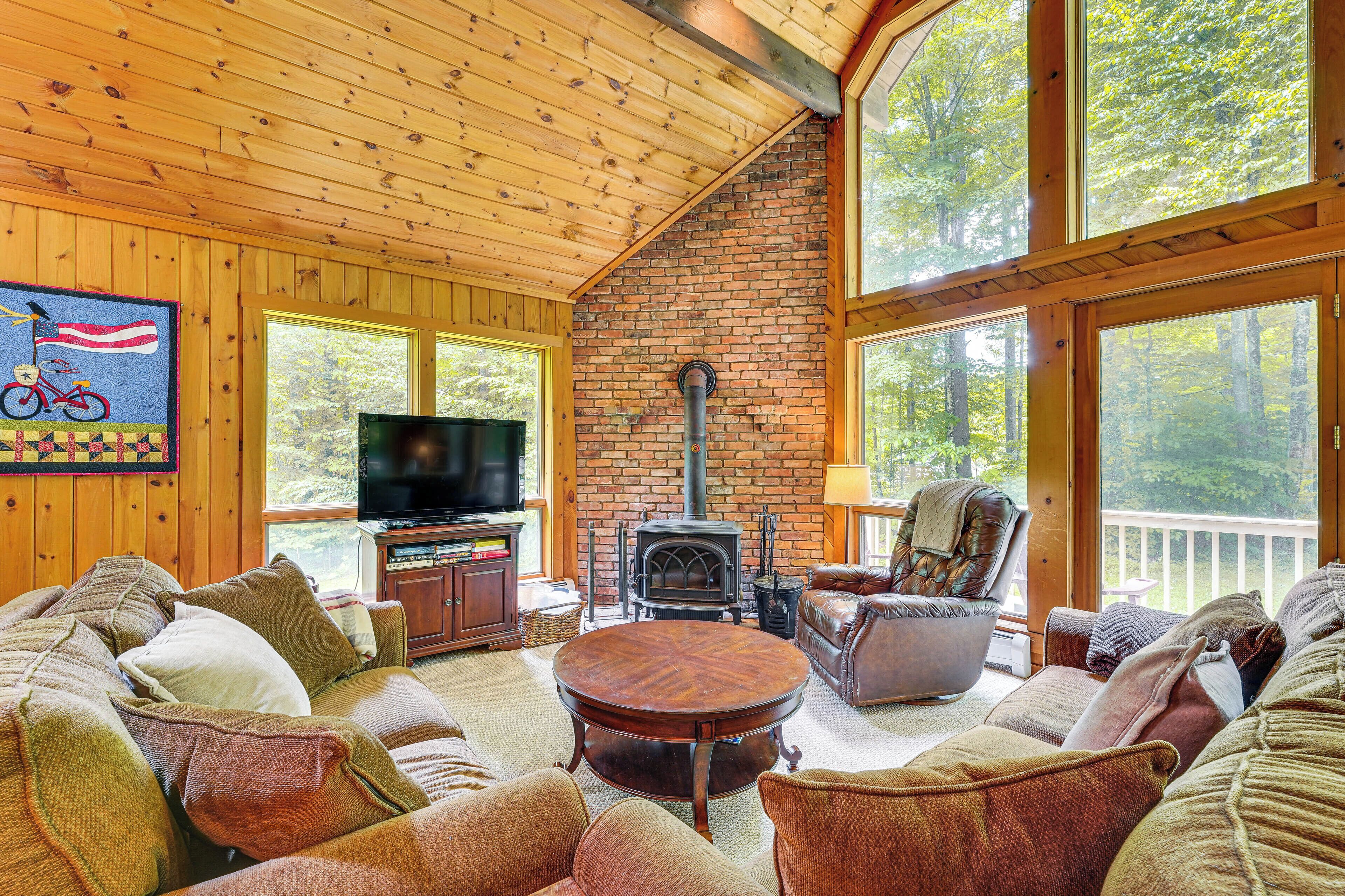 A cozy cabin interior with a brick fireplace, wooden ceiling, and large windows offering a view of the forest.