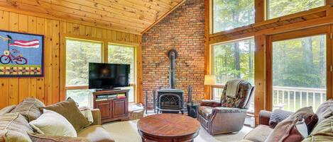 A cozy cabin interior with a brick fireplace, wooden ceiling, and large windows offering a view of the forest.