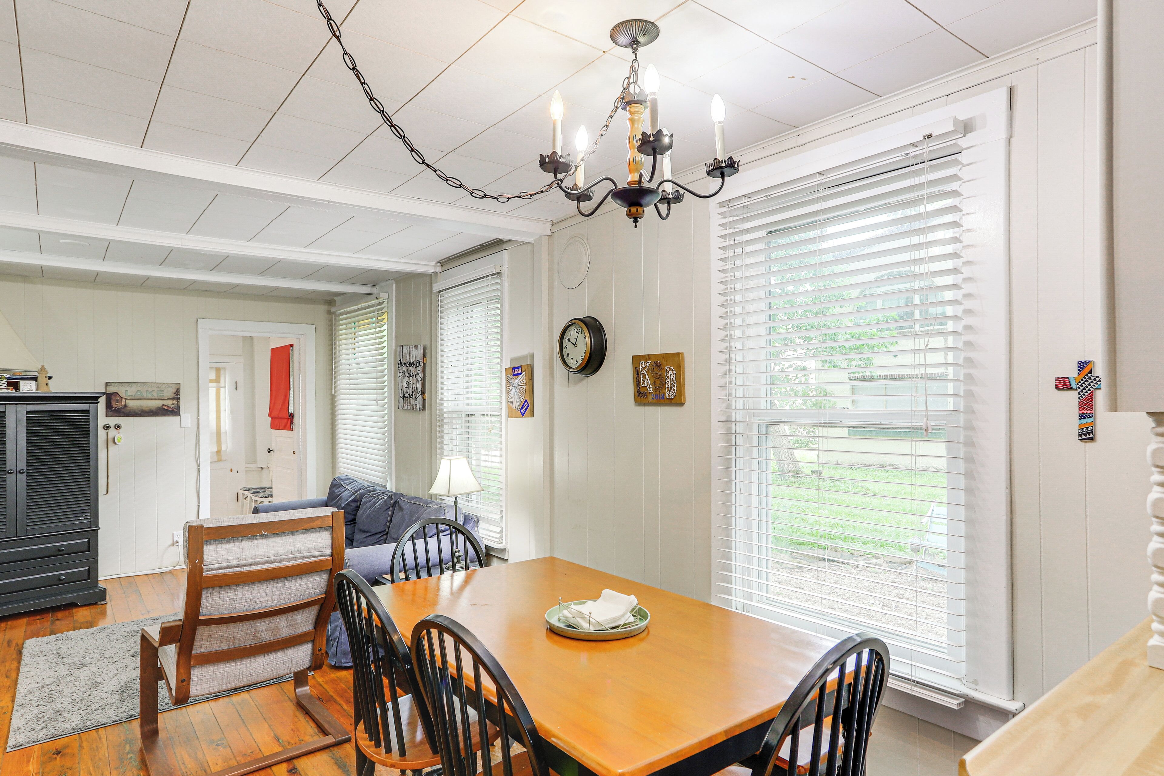 A dining area with a wooden table, chairs, and a chandelier. There is a clock on the wall and a window with blinds.