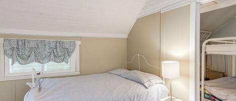 A bedroom with a bunk bed, a bedside table with a lamp, a window with a lace curtain, and a bed with striped bedding.