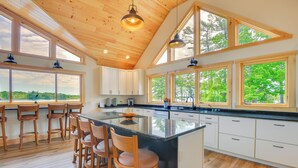 Une cuisine spacieuse avec des meubles en bois, un grand îlot central doté de tabourets, et de grandes fenêtres offrant une vue sur l’extérieur.
