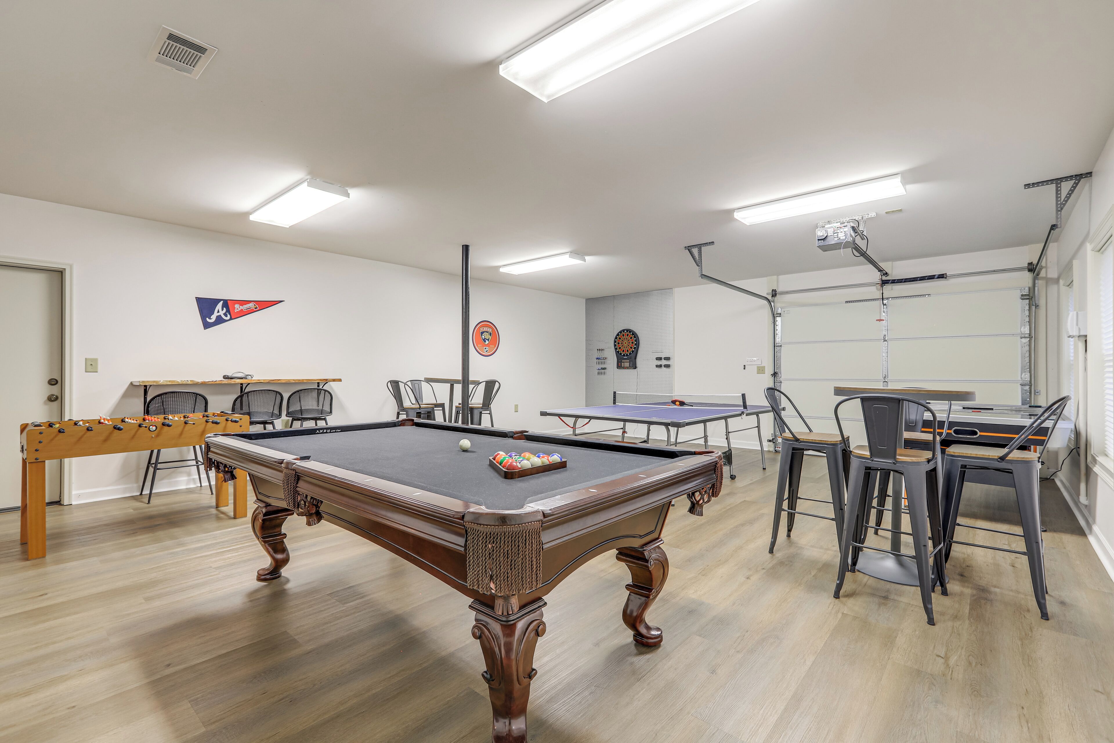 A pool table with a cue ball and a rack of balls, surrounded by chairs and a ping pong table in a well-lit room with white walls and wooden flooring.