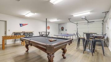 A pool table with a cue ball and a rack of balls, surrounded by chairs and a ping pong table in a well-lit room with white walls and wooden flooring.
