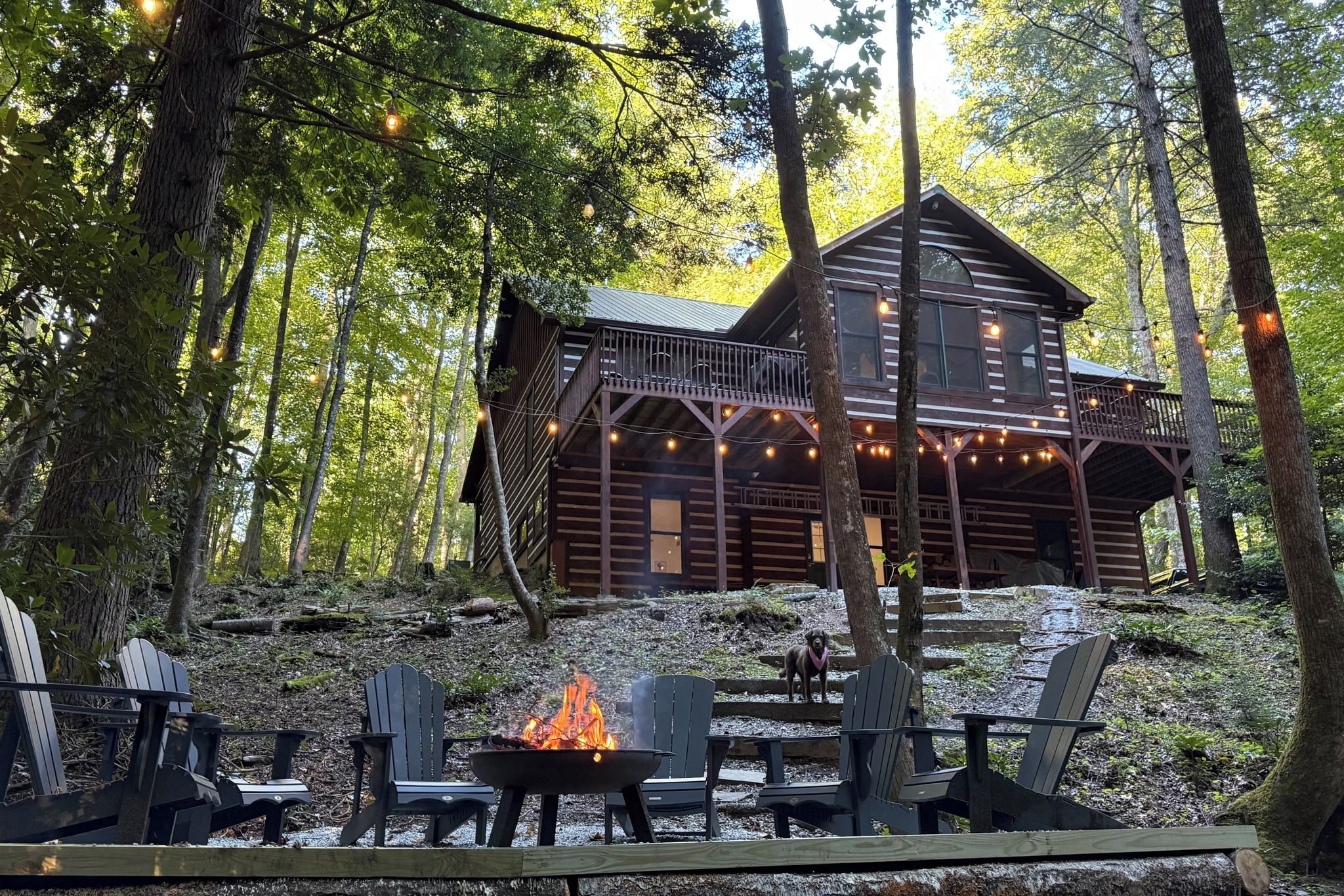 A cabin with a deck, surrounded by trees, and a fire pit with chairs in front.