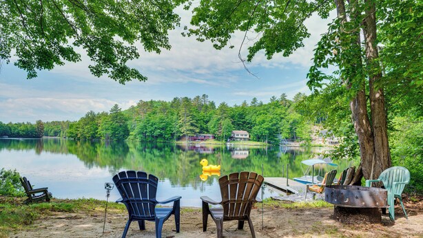 A lakeside setting with chairs and a fire pit, surrounded by trees and a view of a lake and distant buildings. - Denmark Waterfront Cabin w/ Dock & Charcoal Grill! (Denmark)