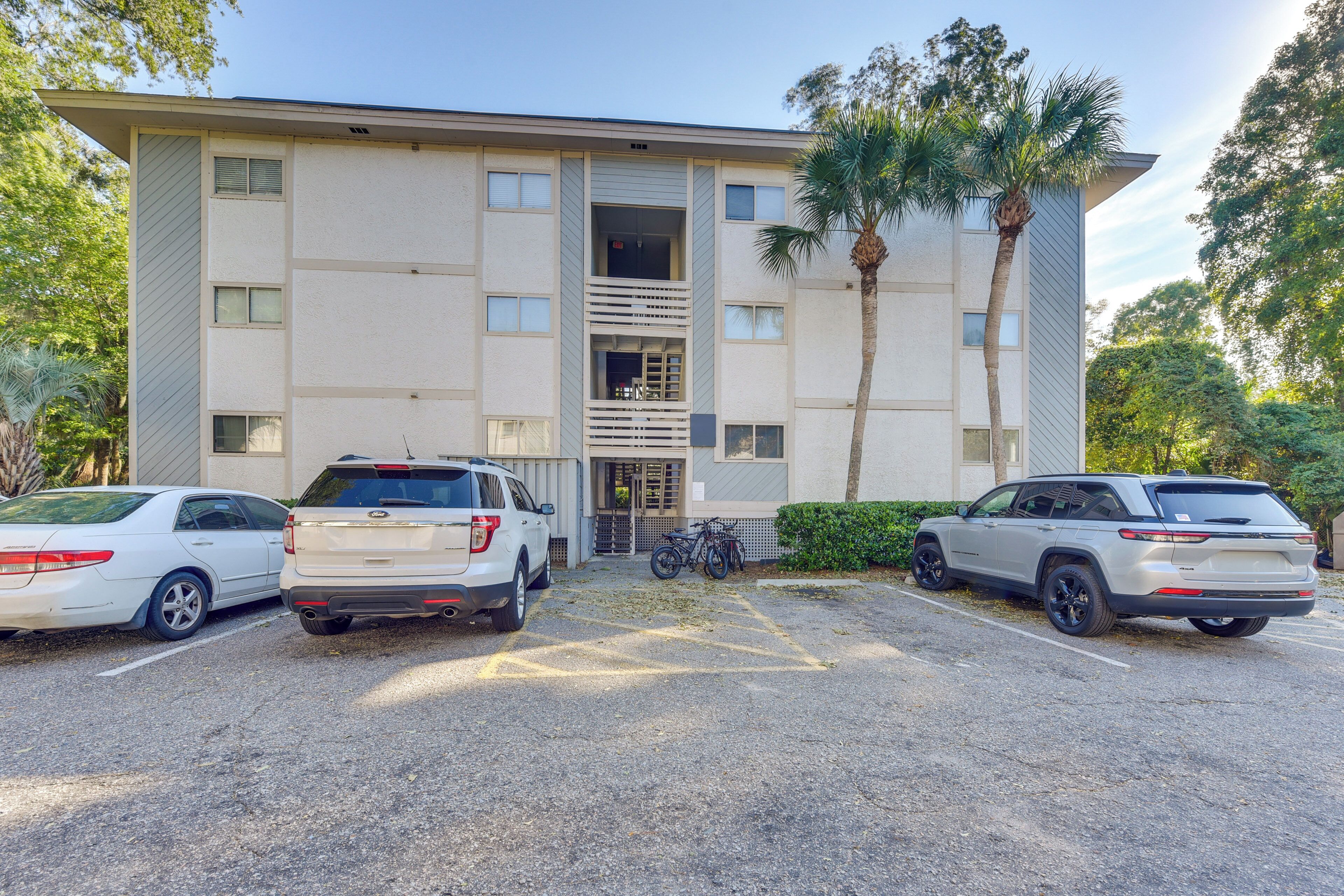 A parking lot with three vehicles, a building with a central entrance, and a palm tree.