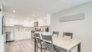 A modern kitchen with white cabinets, a stainless steel refrigerator, a microwave, and a dishwasher. There is a dining table with four chairs and a 'Welcome' sign on the wall.