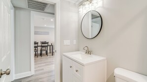 A bathroom with a white sink, a round mirror, and a toilet. There is a door leading to a dining area with a table and chairs.