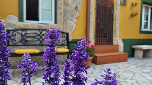 A stone-paved entrance with purple flowers in the foreground. A yellow building with a green window frame and a black metal gate is visible in the background. - La Casita A de Naval in the Rías Baixas, Pontevedra (activities for customers)  (Pontevedra)