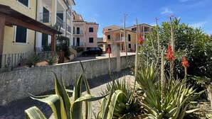 A residential street with multi-story buildings, a car parked on the side, and a garden with large plants and red flowers. - Affittimoderni Valledoria - Sa Turritta (Valledoria)