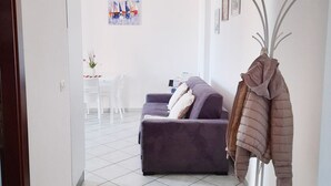 A living room with a sofa, a coat rack with a jacket, and a dining area in the background. - Affittimoderni Valledoria - Sa Turritta (Valledoria)