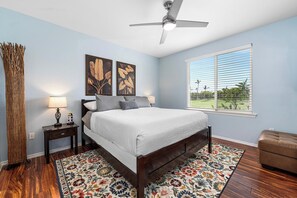 Un dormitorio con cama, mesita de noche, lámpara, ventilador de techo y ventana con persianas, y vista a zonas verdes al exterior.