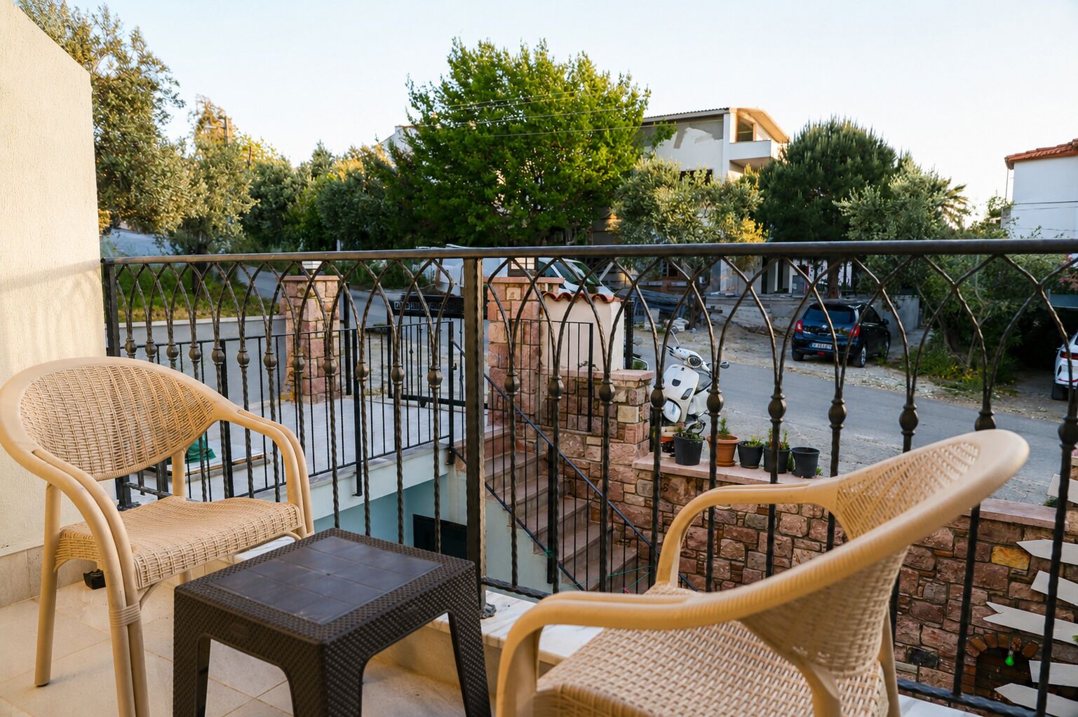 A balcony with wicker chairs and a small table, a stone wall, and a view of parked cars and greenery.