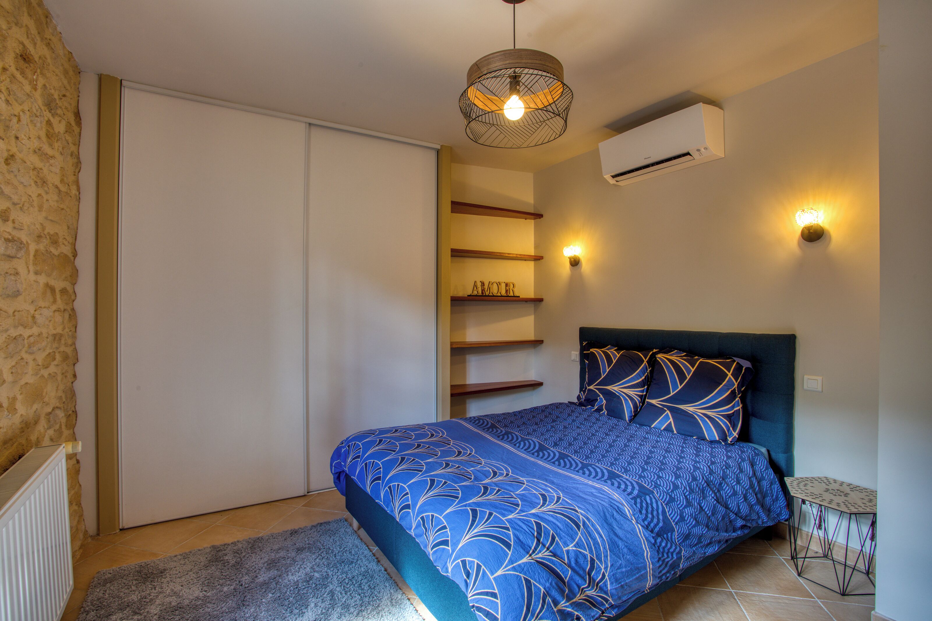Une chambre à coucher comprenant un lit, un climatiseur fixé au mur, un luminaire au plafond et une petite table avec une chaise.