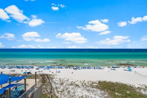 A beach with clear turquoise water, white sand, and a view of the ocean. There are blue umbrellas and lounge chairs on the beach.