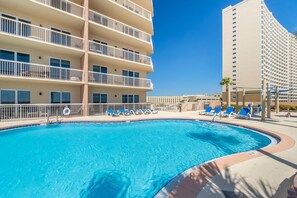 A swimming pool with lounge chairs, a building with balconies, and a clear blue sky.