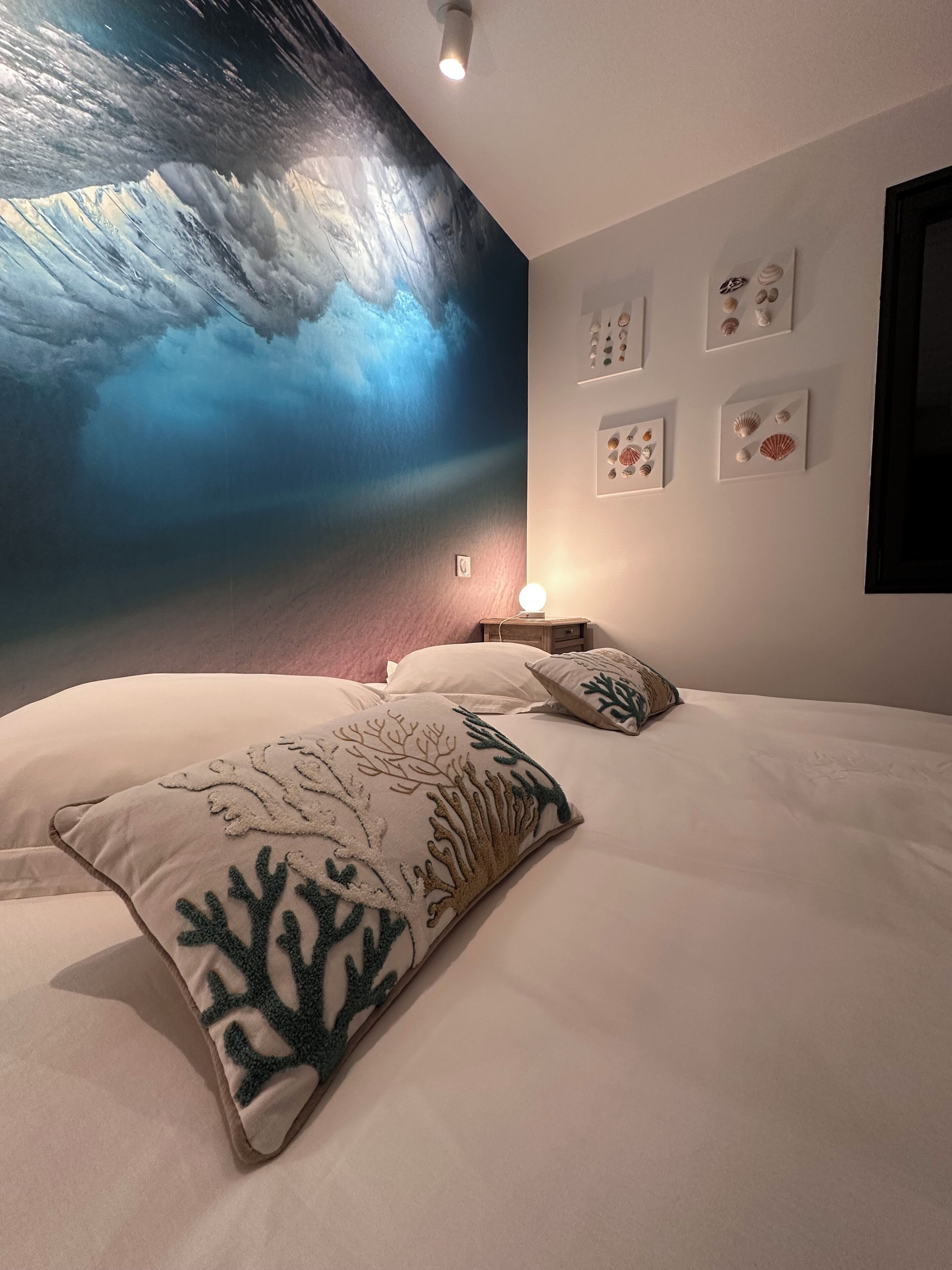 A bedroom with a large mural of a wave, a bed with two decorative pillows, and several framed pictures on the wall.