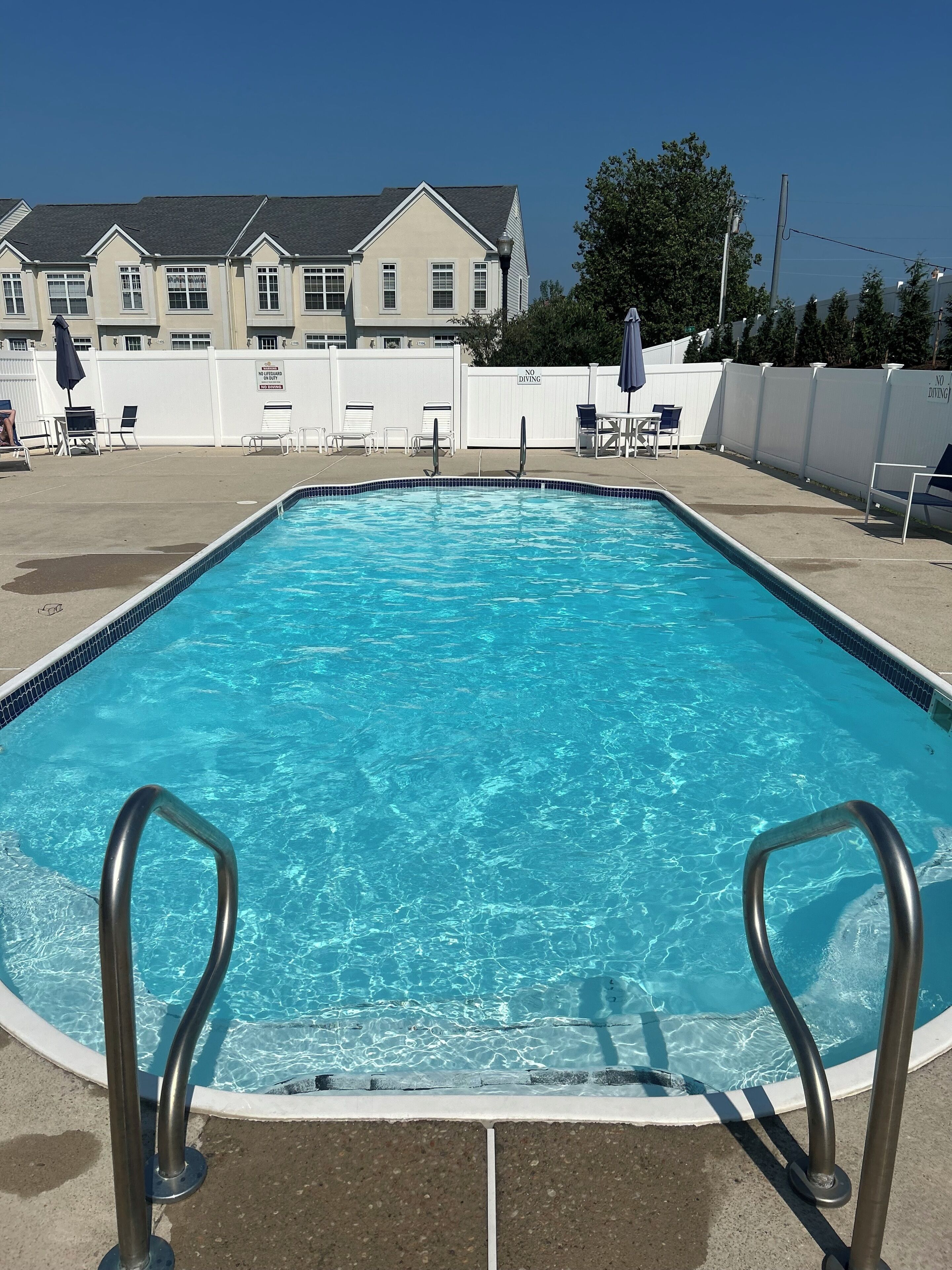 A rectangular swimming pool with clear blue water, surrounded by a white fence, and a row of lounge chairs and umbrellas.