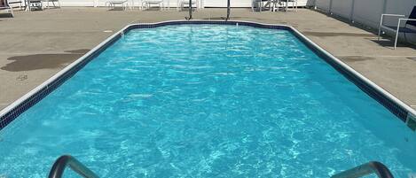 A rectangular swimming pool with clear blue water, surrounded by a white fence, and a row of lounge chairs and umbrellas.