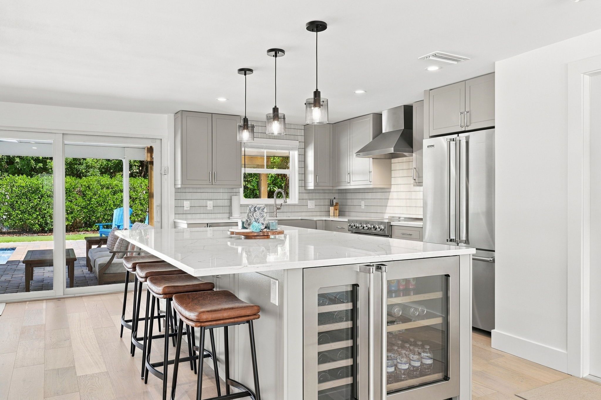A modern kitchen with a large island, bar stools, stainless steel appliances, and a sliding glass door leading to an outdoor area.