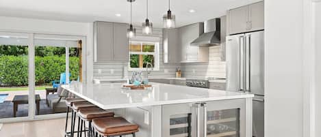 A modern kitchen with a large island, bar stools, stainless steel appliances, and a sliding glass door leading to an outdoor area.